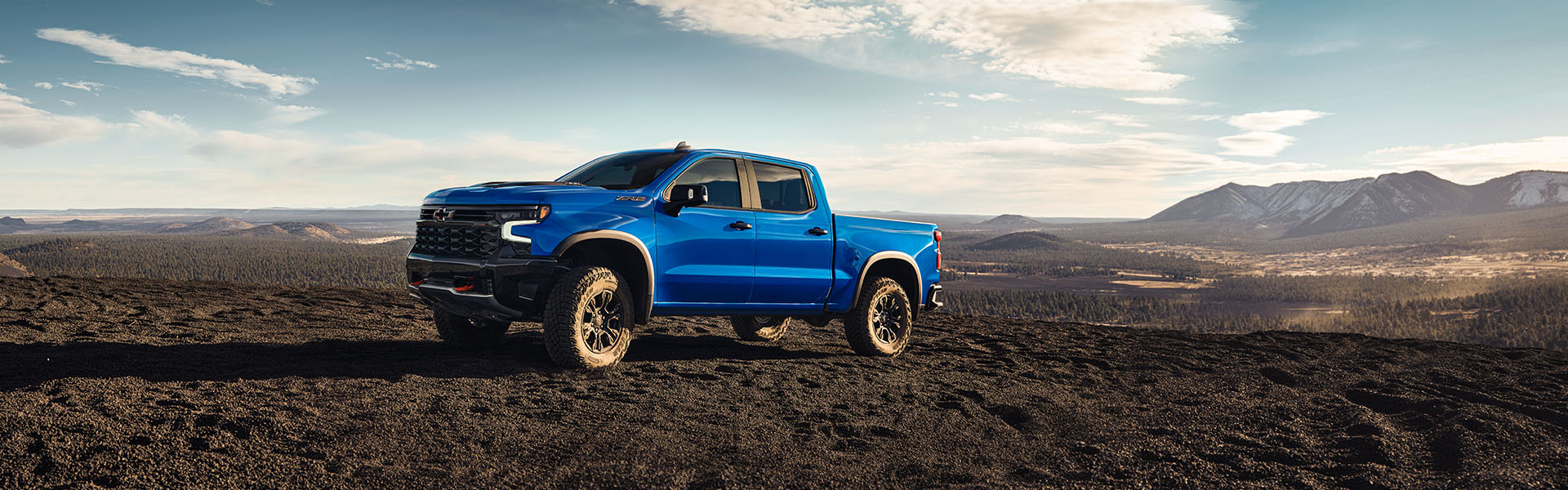 Blue 2025 Chevrolet Silverado 1500 parked on rugged off-road terrain with mountains and wide-open desert landscape in the background