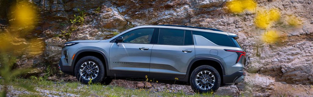 Side profile of a grey 2026 Chevy Traverse parked on a grassy slope against a rugged rock cliff.