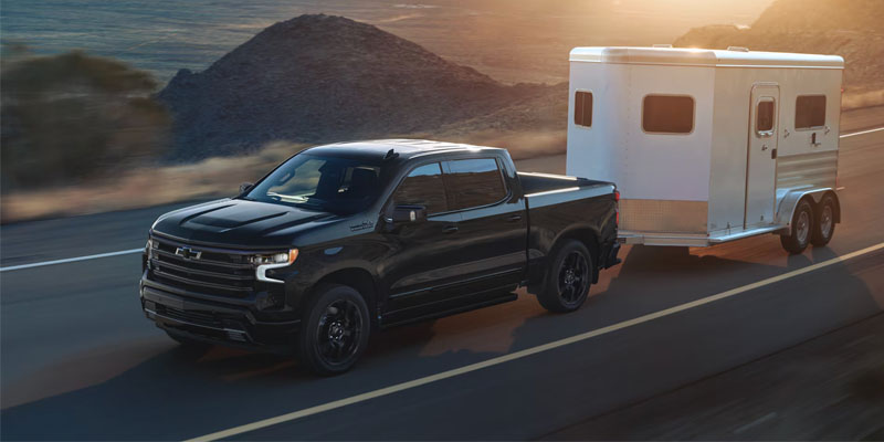 A sleek black pickup truck Chevrolet-Silverado-1500 tows a white trailer on a sunlit mountain road. The setting sun casts a warm glow, emphasizing the rugged, adventurous tone.