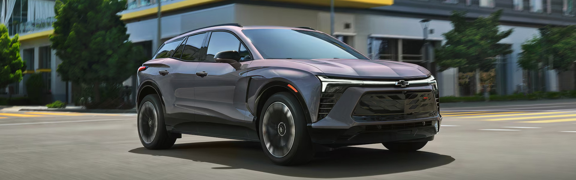2026 Chevrolet Blazer RS midsize SUV in gray, driving through an urban street with a sleek sporty design, black grille, LED headlights, and modern city buildings in the background.